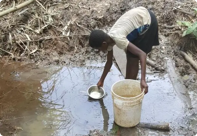 A person collects water from a muddy puddle using a bowl, pouring it into a white bucket, in a rural, undeveloped area.