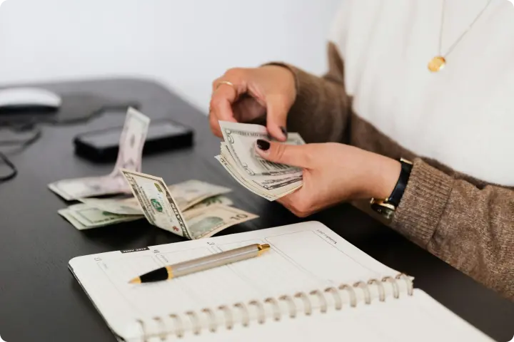 Person counting dollar bills at a desk with a notebook, pen, and smartphone nearby.