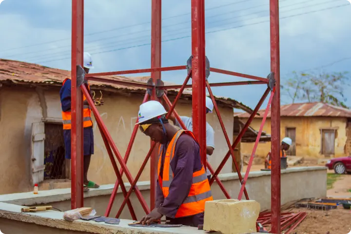 Construction workers wearing helmets and orange vests work on a building site with red metal beams and rustic structures in the background.