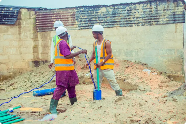 Construction workers in safety vests and helmets operate equipment on a sandy site near a corrugated metal and brick wall.