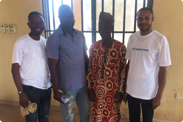 Four men standing together indoors, three wearing casual shirts and one in traditional patterned attire, smiling at the camera.