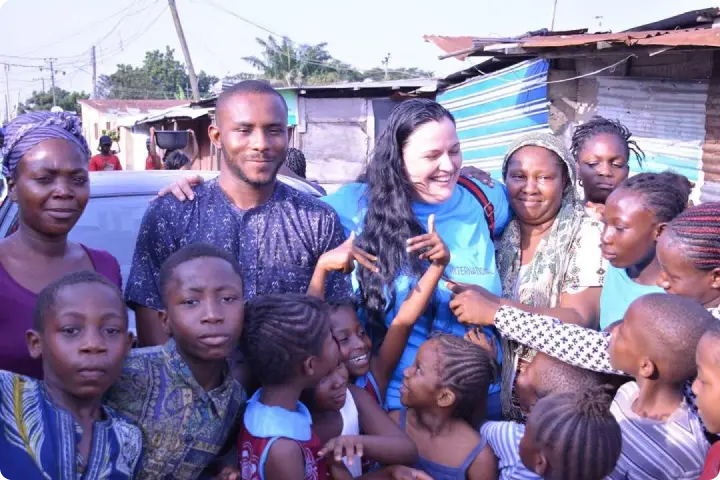 A group of smiling adults and children gather closely in an outdoor setting, with makeshift structures in the background.