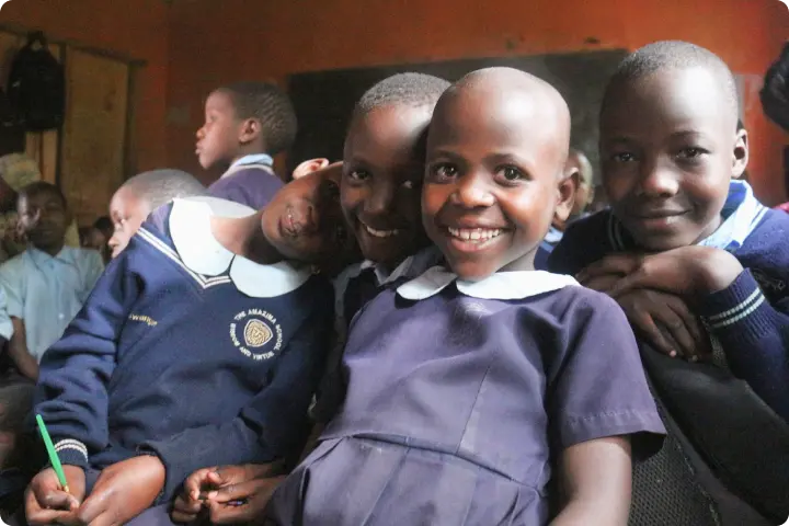 Four children in school uniforms smiling and posing together in a classroom, with one holding a green pen.