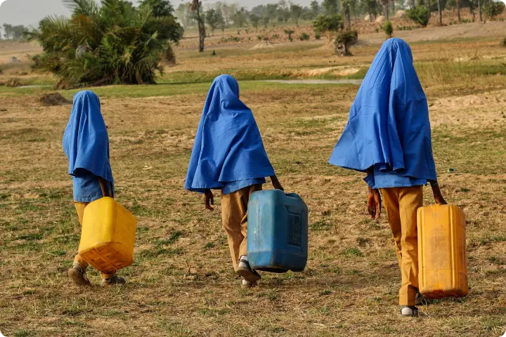 Three children walk across a dry field, carrying large water containers and covered with blue cloths.