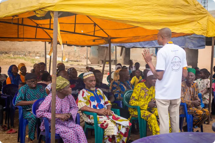A group of people seated under a yellow tent, listening to a standing speaker in a white shirt, in an outdoor setting.