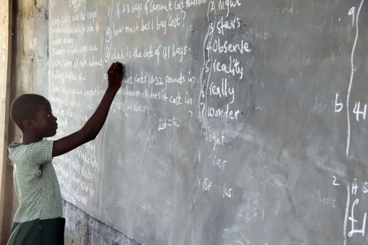 A student writes on a chalkboard covered with text in a classroom, focusing intently on the task.