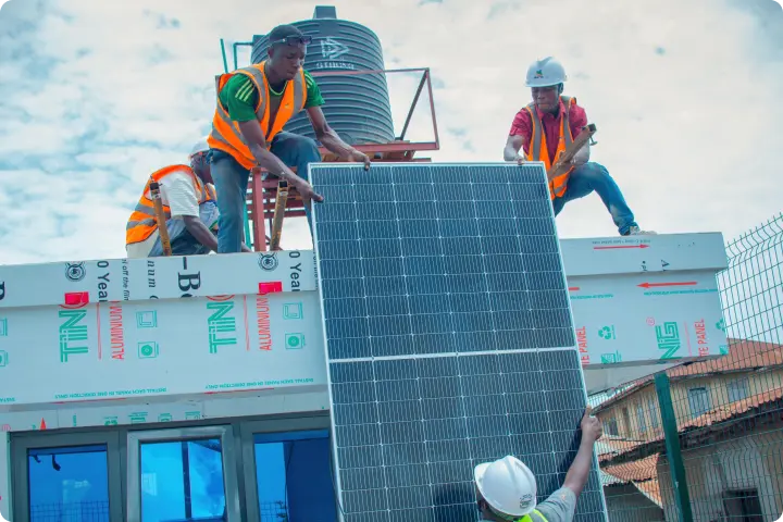 Workers in safety gear install a large solar panel on a rooftop, with a water tank visible in the background under a partly cloudy sky.