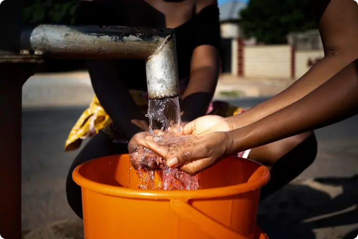 Two people collect water in cupped hands from a metal spout, with an orange bucket below to catch the overflow.