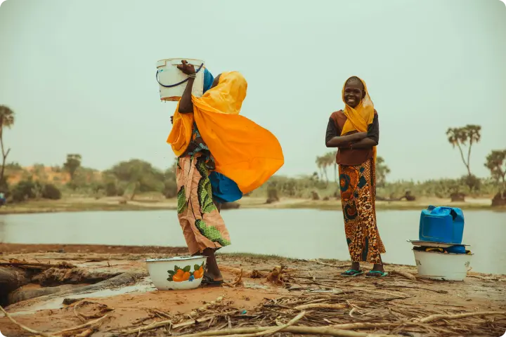 Two people by a river, one pouring water from a bucket, both wearing colorful clothing, with trees in the background.