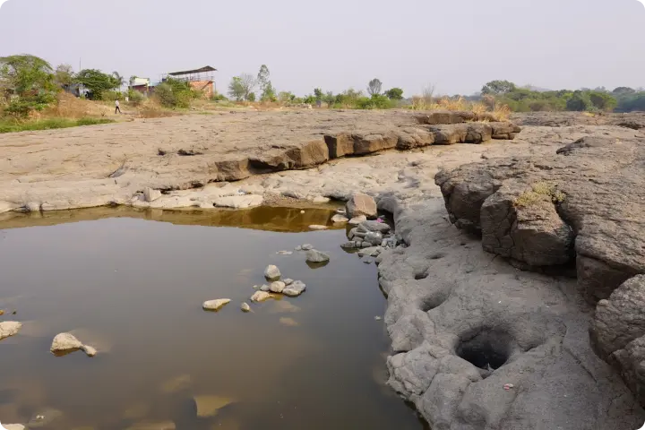 Rocky terrain with shallow water pools and scattered stones. Sparse vegetation and a small structure are visible in the background.