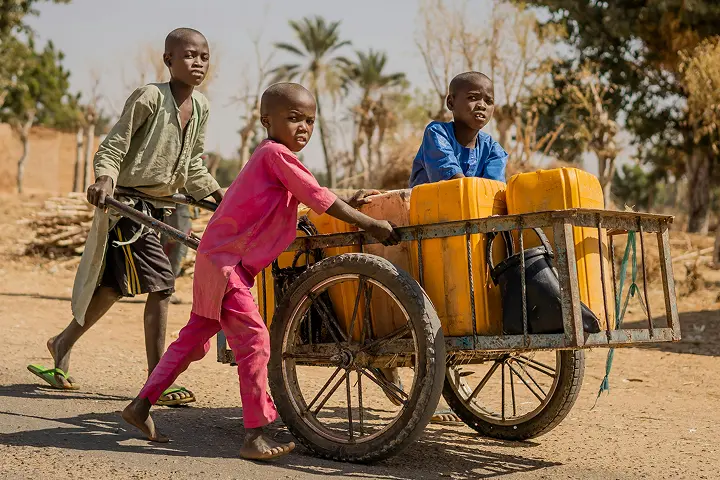 Three children push a cart loaded with yellow jerry cans along a dirt path, with trees and dry vegetation in the background.