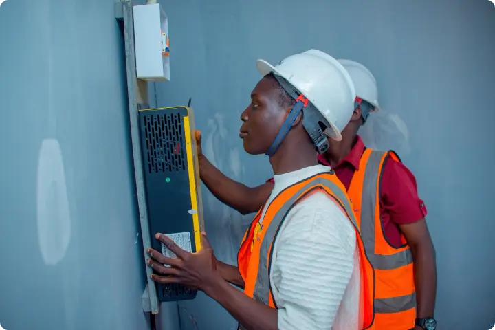 Two workers in safety gear and helmets install equipment on a wall. One holds a device while the other adjusts a panel.