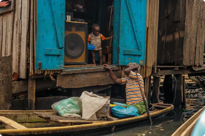 A person in a striped shirt sits in a canoe on a waterway, looking up. A child stands in a doorway of a blue wooden building above.