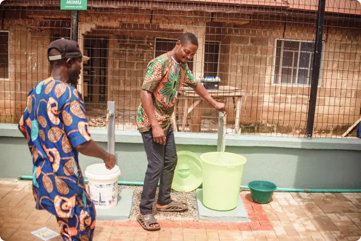Two men in colorful shirts collect water from a communal tap into large buckets outside a building with barred windows.