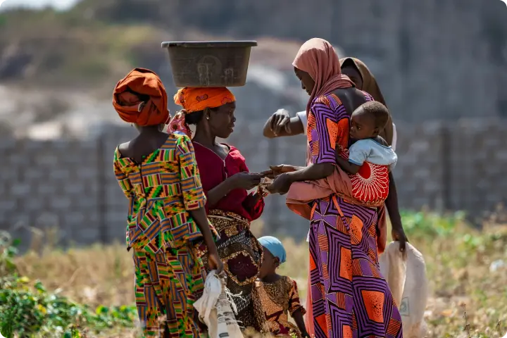 A group of women in colorful clothing, one carrying a baby and another balancing a basket on her head, gather outdoors in a rural setting.