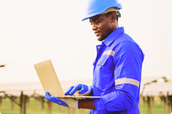 Man in blue work attire and helmet using a laptop outdoors near solar panels.
