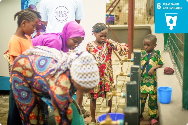 Group of children and adults gather around a water tap station, filling containers. A "Clean Water and Sanitation" banner is visible.