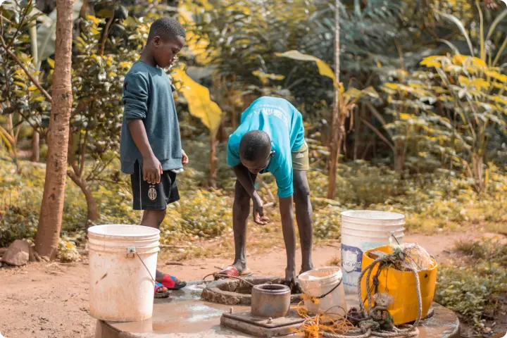 Two boys gather water at an outdoor pump, surrounded by buckets and lush greenery.