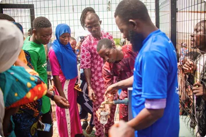 A group of people gather around a man pouring water from a bottle, with some wearing colorful clothing and headscarves, near a metal fence.