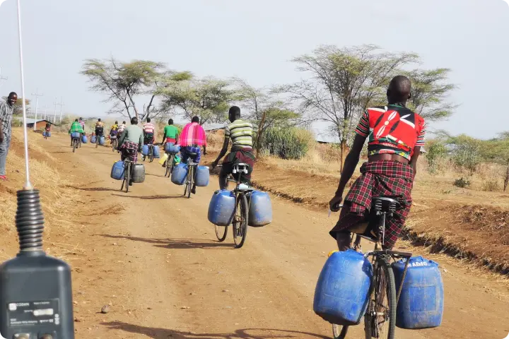 Cyclists on a dirt road carry large blue containers. Sparse trees and dry grass line the path under a clear sky.