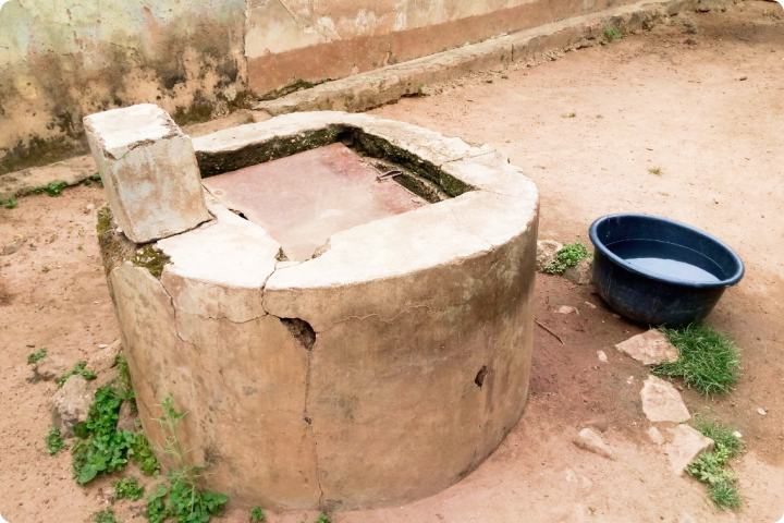 A weathered, cracked concrete well with a rectangular cover, next to a blue plastic bucket partially filled with water, on a dirt surface.