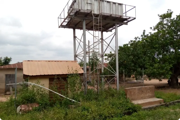 Elevated metal water tank on a tall structure beside small buildings and trees, set against a cloudy sky.