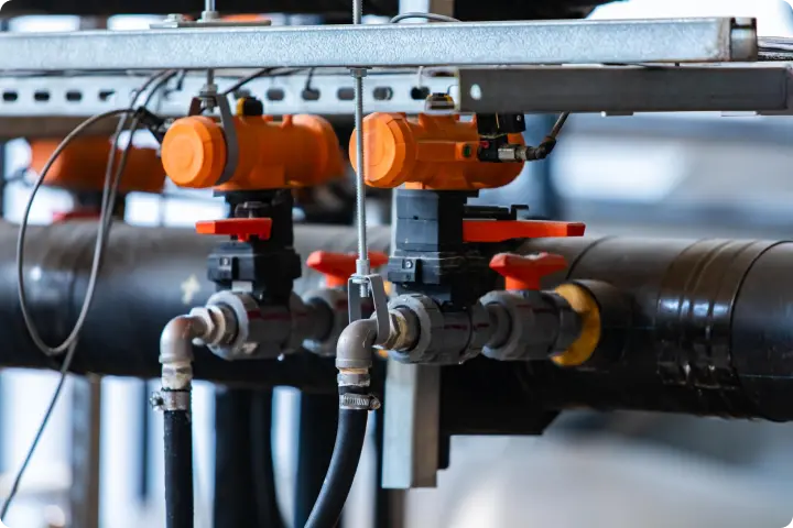 Industrial piping system with orange valve actuators and red-handled valves, connected by black pipes and metal supports.