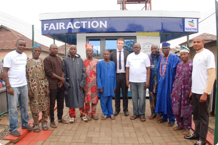 A group of people, including men in traditional attire and others in white shirts, stand in front of a building labeled "Fairaction."