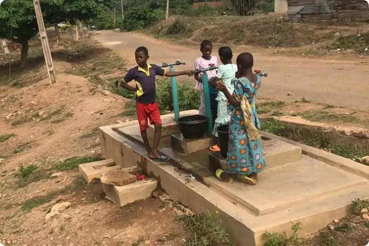 Four children gather around a water pump on a dirt path, filling a black bucket. Trees and a building are visible in the background.