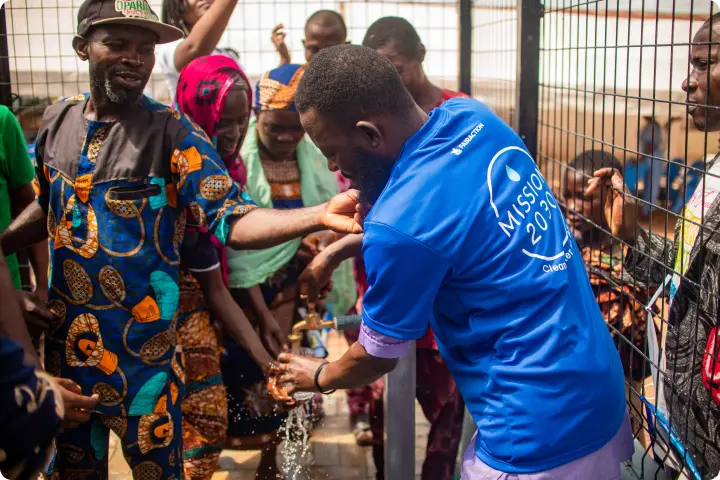 A group of people gather around a man in a blue shirt as he helps them wash hands at an outdoor water tap station.