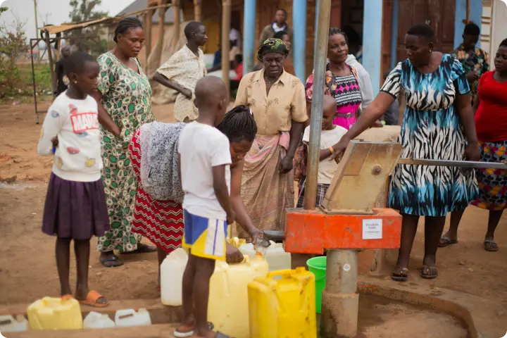 People gather around a communal water pump, filling containers with water. A child and several adults are actively engaged in the task.