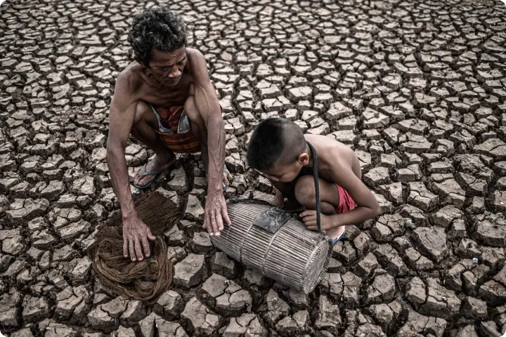 An elderly man and a boy sit on cracked, dry earth, examining fishing nets and a basket, symbolizing drought and livelihood challenges.