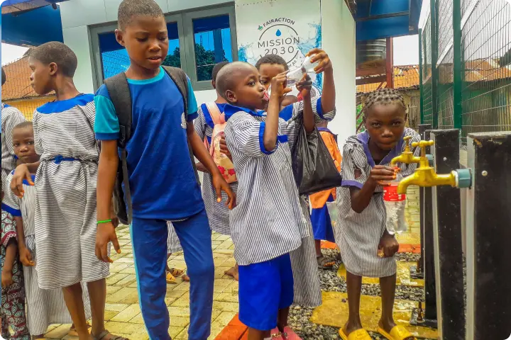 Children in school uniforms gather around a water tap, with one boy filling a bottle. A building with a "Mission 2030" sign is in the background.