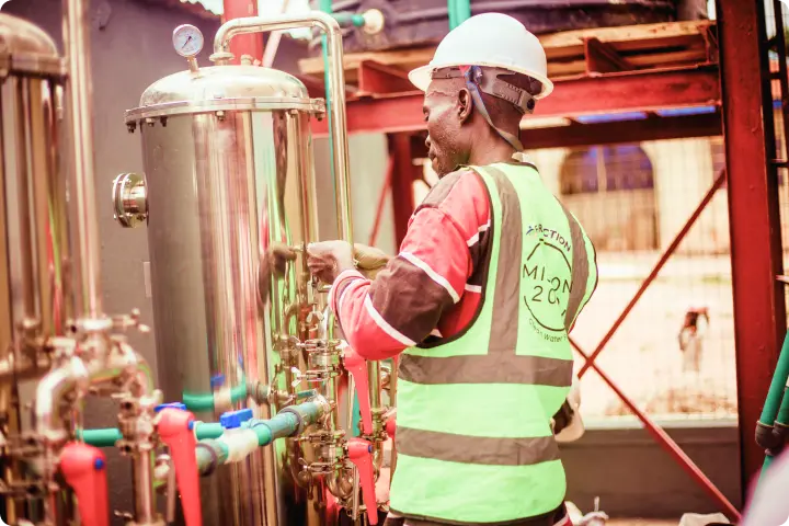 Worker in a safety vest and helmet operates industrial machinery, adjusting valves and gauges in a facility with metal pipes and tanks.