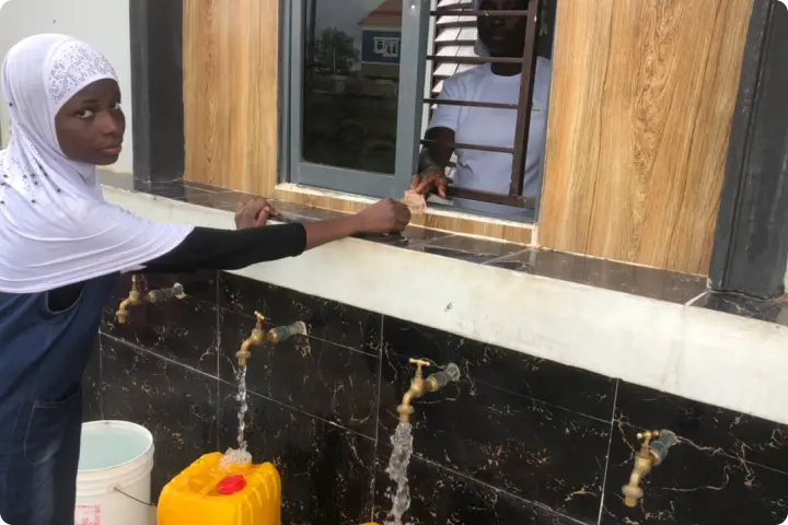 A woman in a white headscarf fills a yellow container with water from a tap outside a building, while another person stands inside by a window.