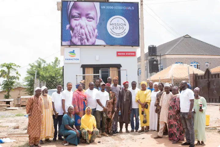 A diverse group of people stands in front of a water station, with a large billboard above promoting UN 2030 Sustainable Development Goals.