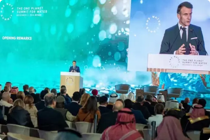A speaker at a podium addresses an audience at the One Planet Summit for Water, with a large screen displaying the event's theme in the background.