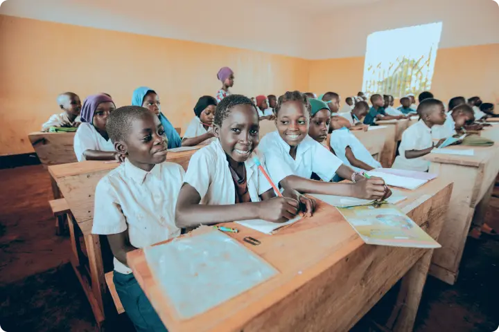 Children in a classroom sitting at wooden desks, smiling and writing in notebooks, with a bright window in the background.