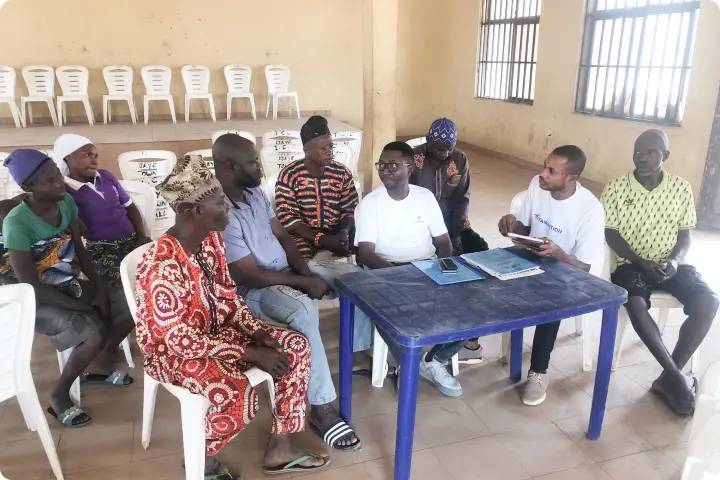 A group of people sitting indoors in a circle, engaged in discussion. Some are seated on plastic chairs around a blue table.