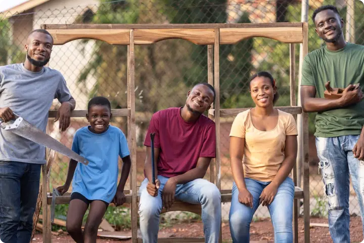 Five people smiling, posing with woodworking tools in an outdoor setting, with a wooden frame and fence in the background.