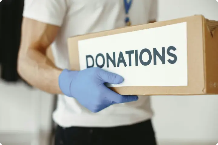 Person wearing blue gloves holding a cardboard box labeled "DONATIONS."