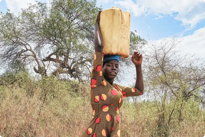 A woman carries a large yellow container on her head, walking through a dry, bushy landscape under a partly cloudy sky.