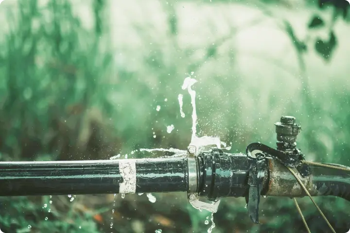 Close-up of a leaking black pipe with water spraying out, set against a blurred green background.