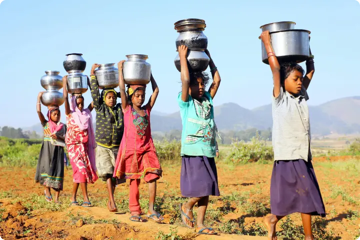 Children walking in a line across a field, balancing metal pots on their heads, with mountains visible in the background.