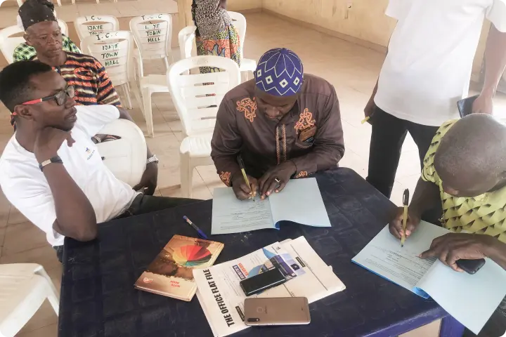 People sitting around a table with documents, notebooks, and phones, engaged in discussion and writing in a room with white chairs.